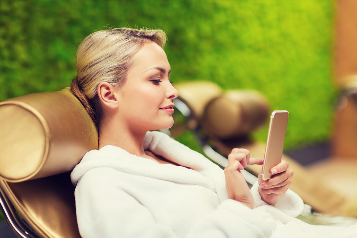 people, beauty, lifestyle, technology and relaxation concept - beautiful young woman in white bath robe with smartphone social networking at spa